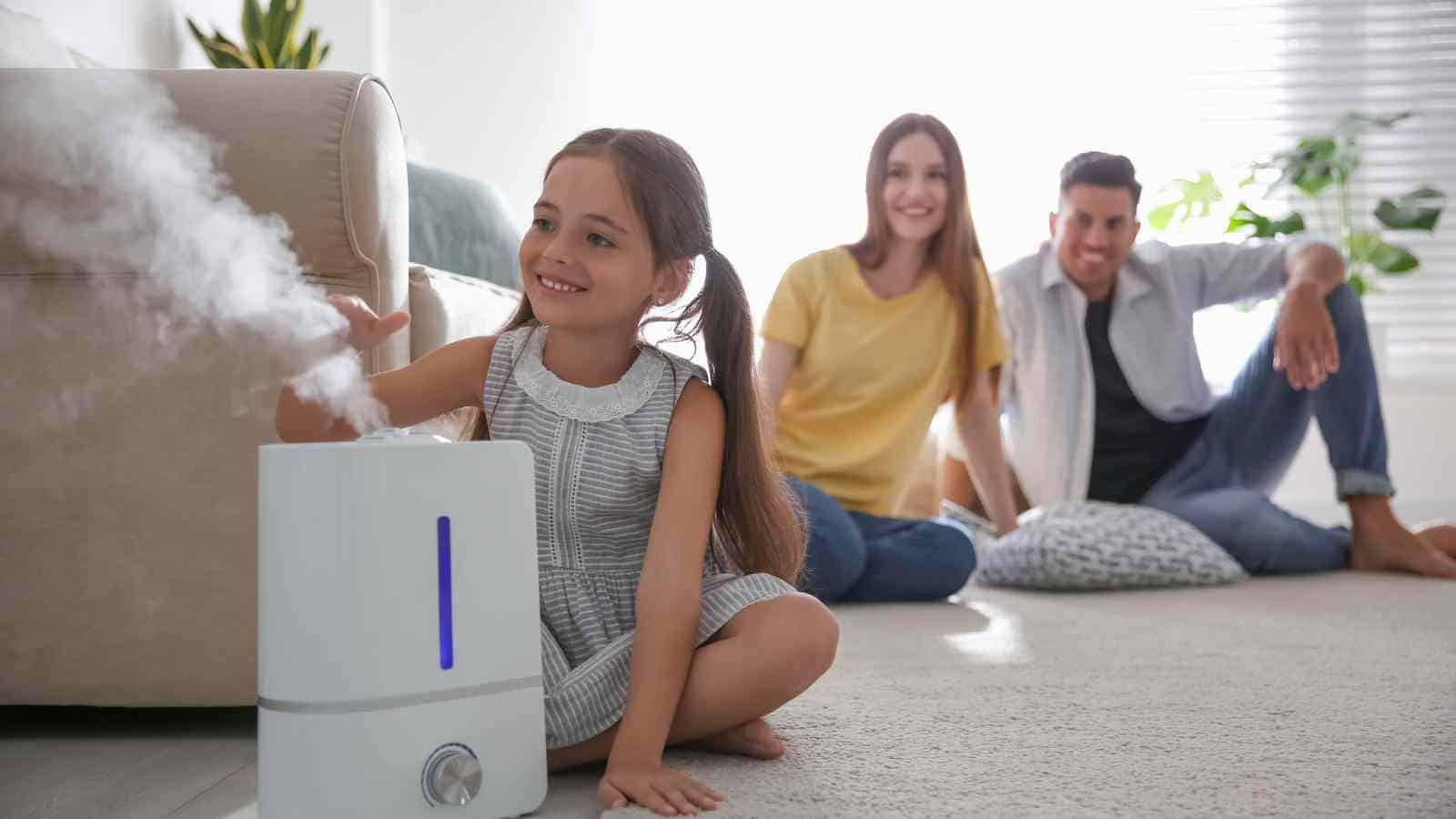 A young girl touches the mist from a humidifier while two adults sit on the floor in the background of a living room.