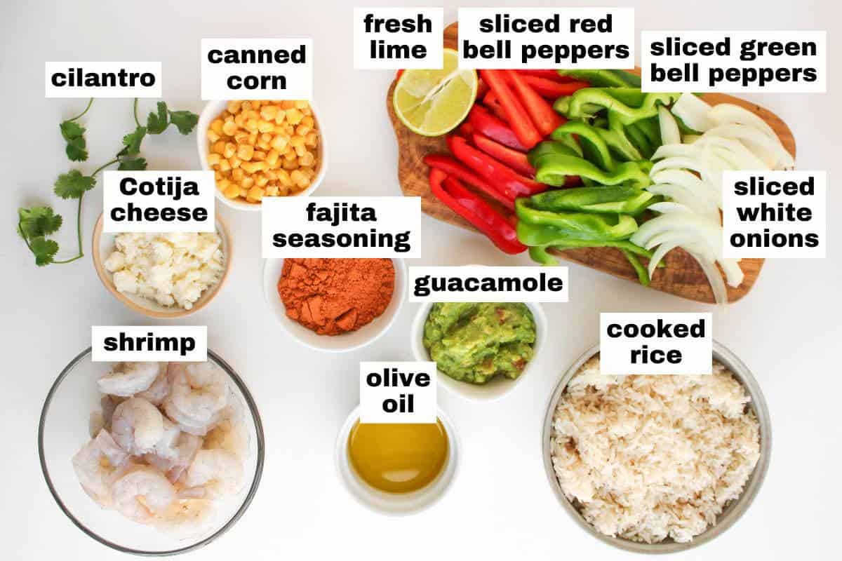 Ingredients for shrimp fajita bowls arranged on a white surface, including shrimp, rice, bell peppers, onions, cilantro, lime, corn, cheese, olive oil, guacamole, and seasoning.