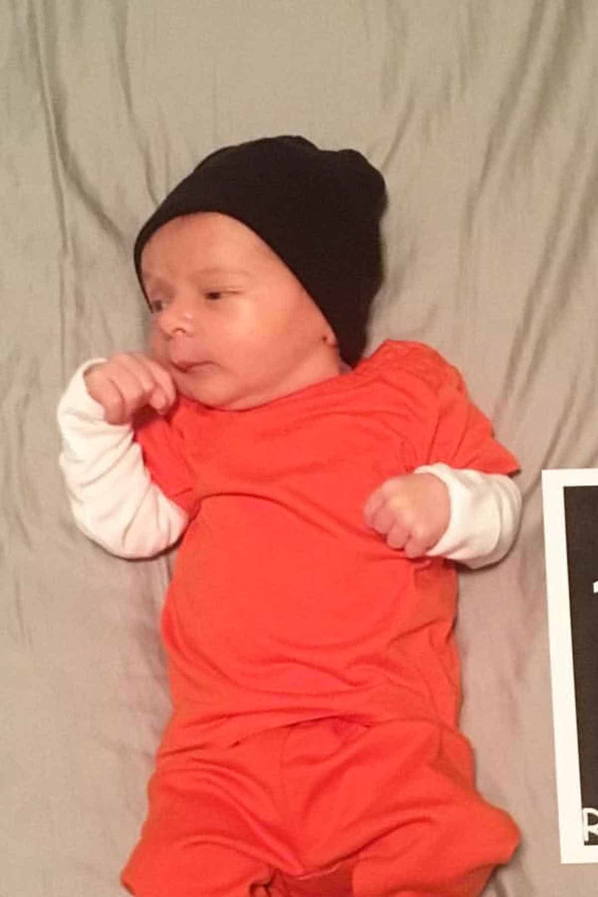 A baby wearing an orange outfit and a black beanie lies on a gray surface next to a partially visible black sign.