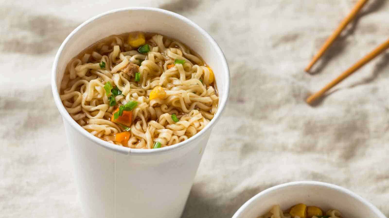 A white paper cup filled with instant ramen noodles, chopped vegetables, and broth sits on a cloth surface with a pair of chopsticks in the background.