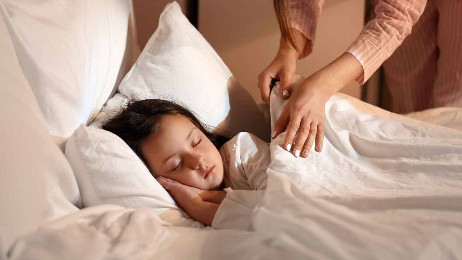 A child sleeps in bed with a hand gently tucking the blanket around them.