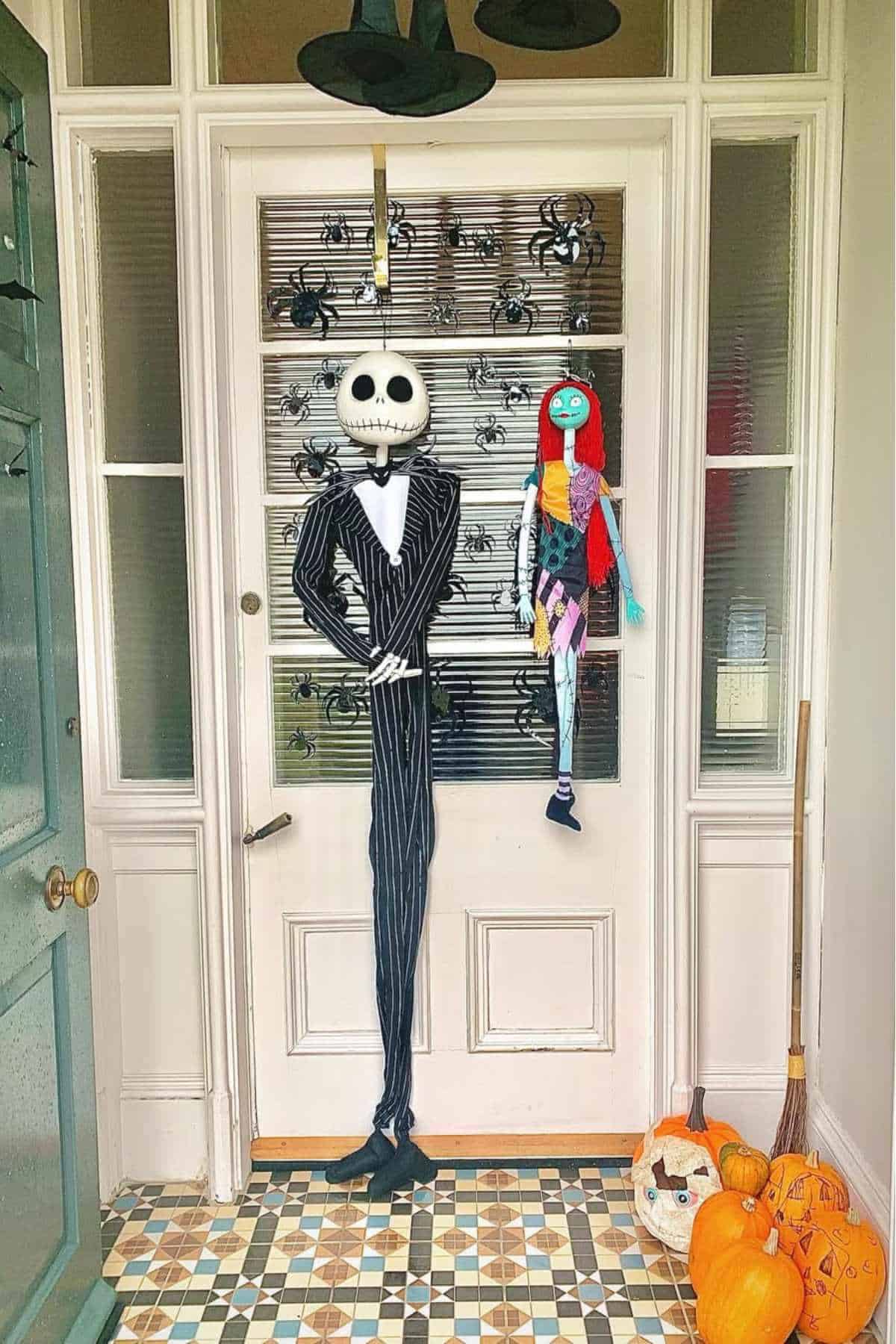 Front door decorated with Jack Skellington and Sally figures, fake spiders on glass, a broom, and pumpkins, including one carved with a face, on patterned tile floor.