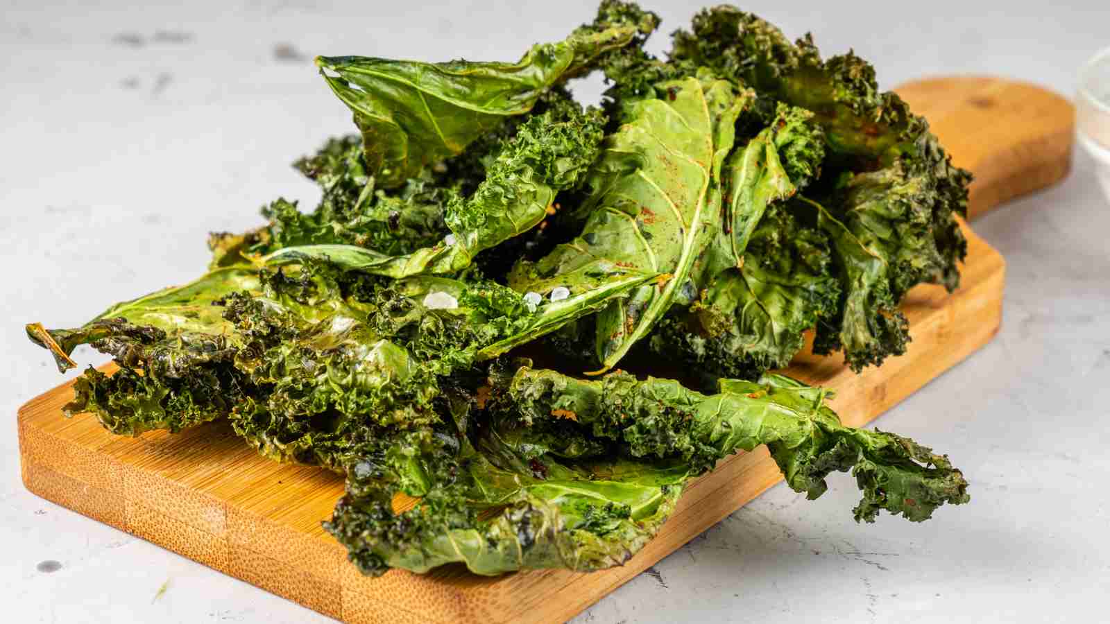 A pile of baked kale chips on a wooden cutting board, with a light-colored background.