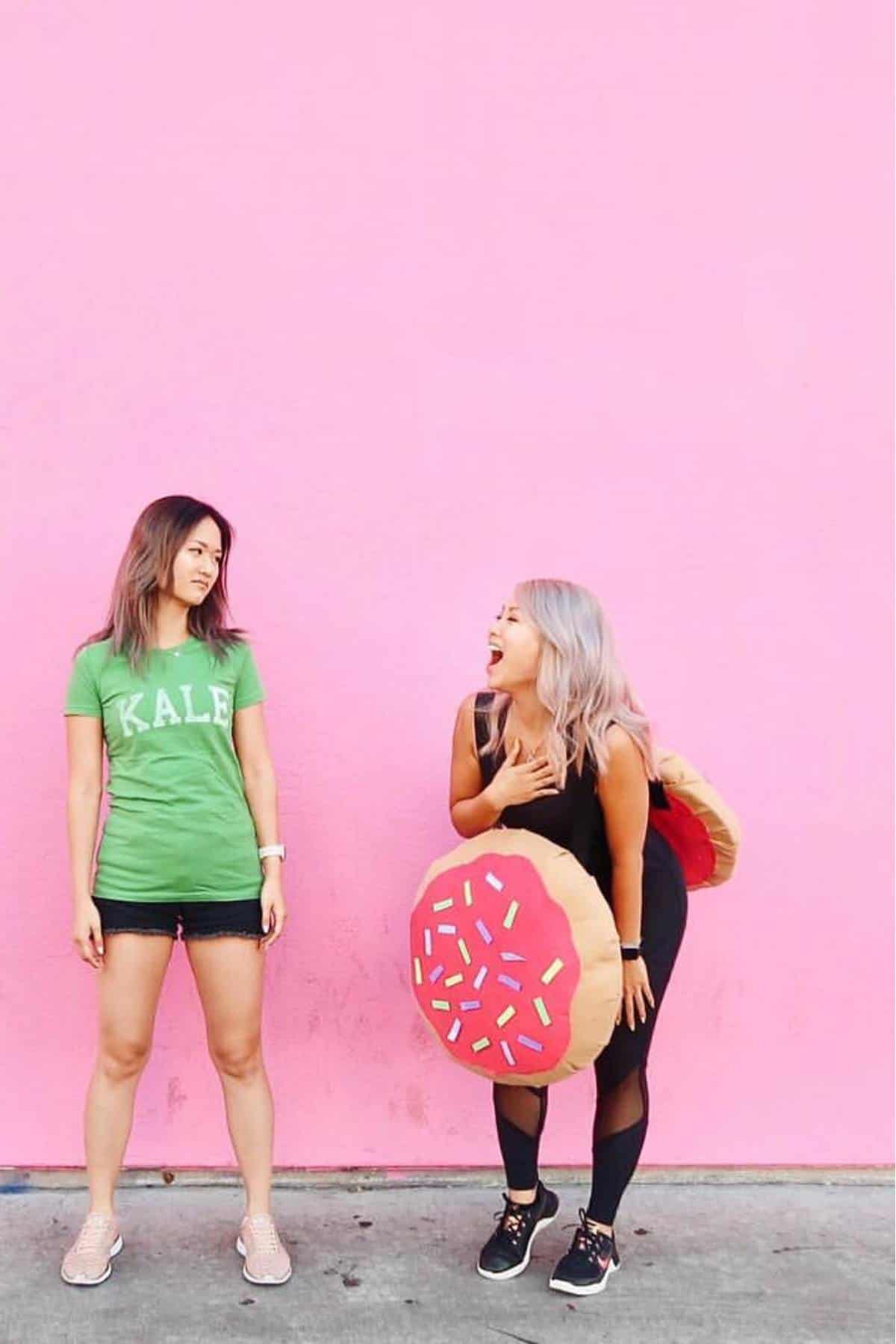 Two women stand in front of a pink wall; one wears a green "Kale" shirt, the other is dressed in a donut costume and laughing.
