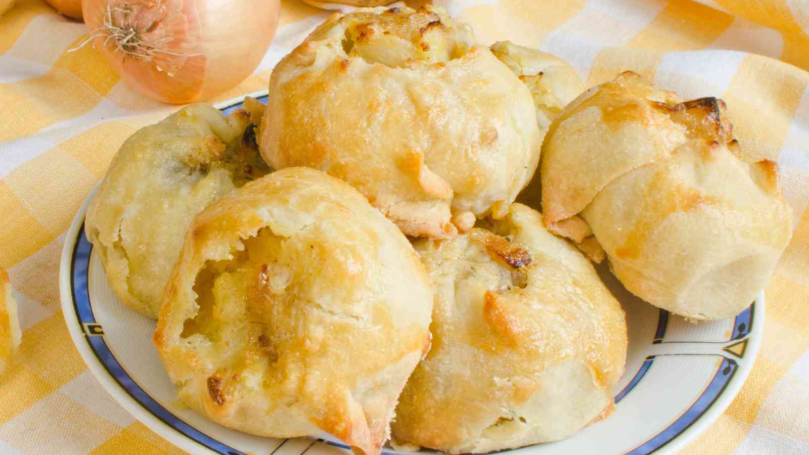A plate with several baked onion rolls sits on a checkered yellow and white cloth, with onions visible in the background.