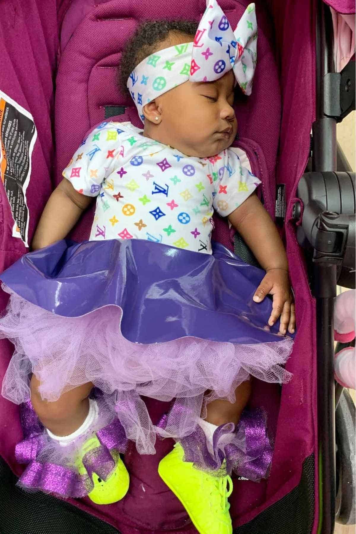 A baby sleeps in a stroller, wearing a colorful headband, a white shirt with multicolored logos, a purple skirt with tulle, and neon yellow shoes.