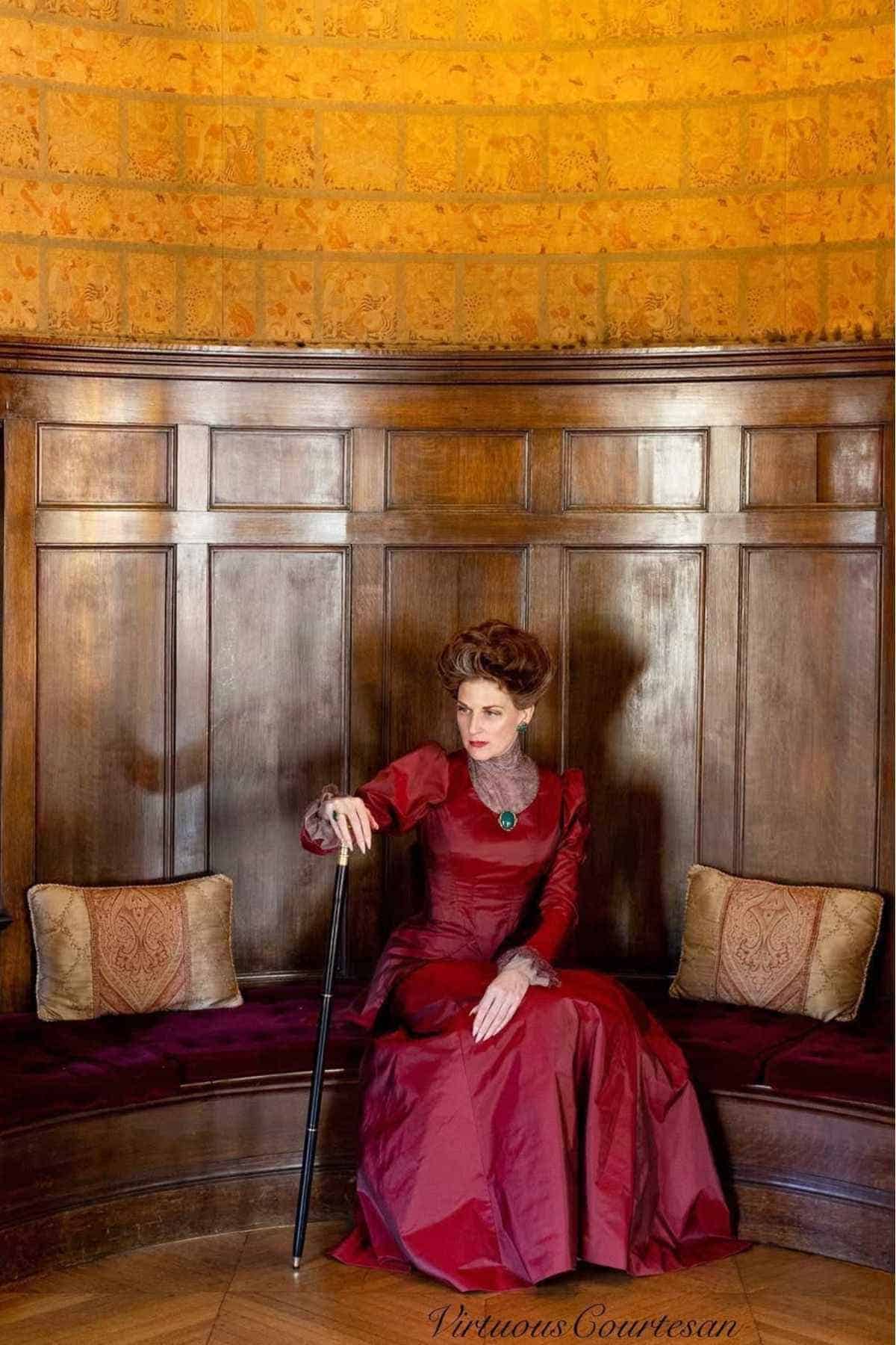 A woman in a long red dress sits on a curved wooden bench, holding a cane, against a wood-paneled wall with two decorative pillows.