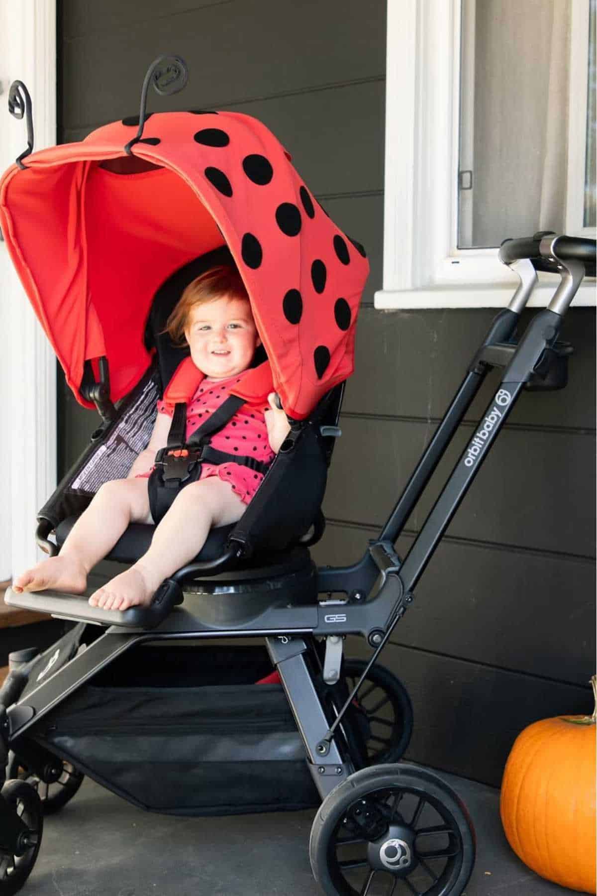 A smiling toddler sits in a stroller with a red and black ladybug canopy, next to a pumpkin on a porch.