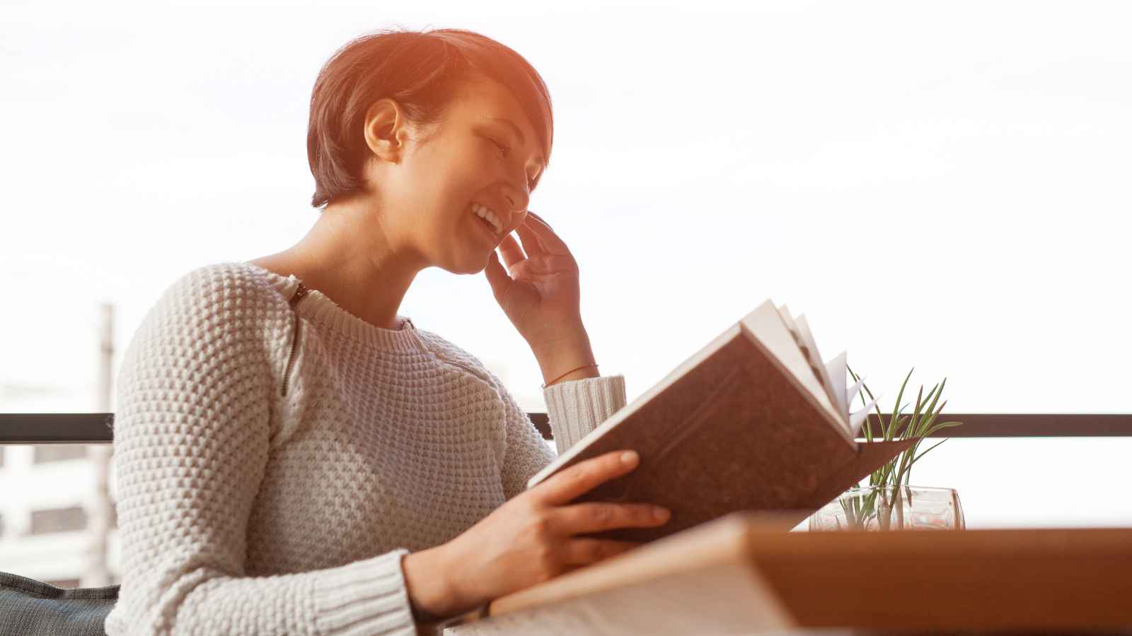 A person wearing a light sweater is sitting indoors, smiling and reading a book at a table with a small plant and a glass beside them.