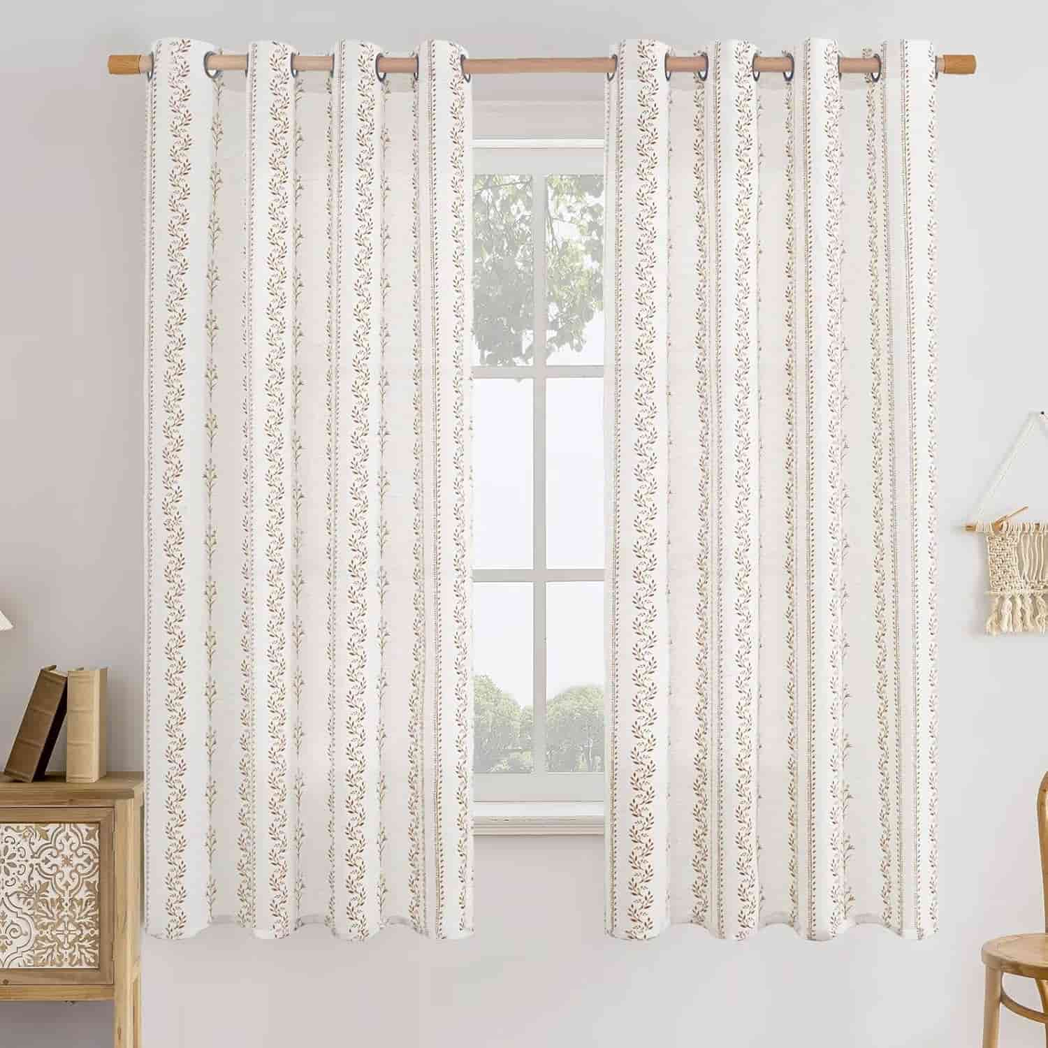 A window with white and beige patterned curtains hanging on a rod, with sunlight filtering through and a partial view of trees outside. A small table with books is on the left.