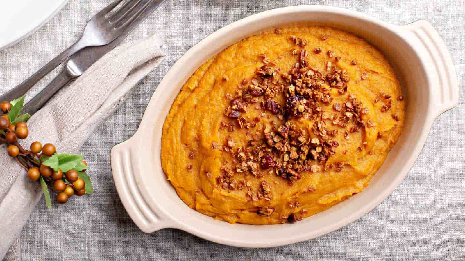 Oval baking dish filled with mashed sweet potatoes, topped with a sprinkling of brown, crunchy granola, placed on a light-colored tablecloth next to cutlery wrapped in a napkin.