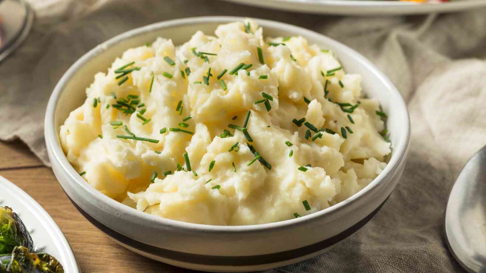 A bowl of mashed potatoes topped with chopped chives sits on a table alongside other dishes.