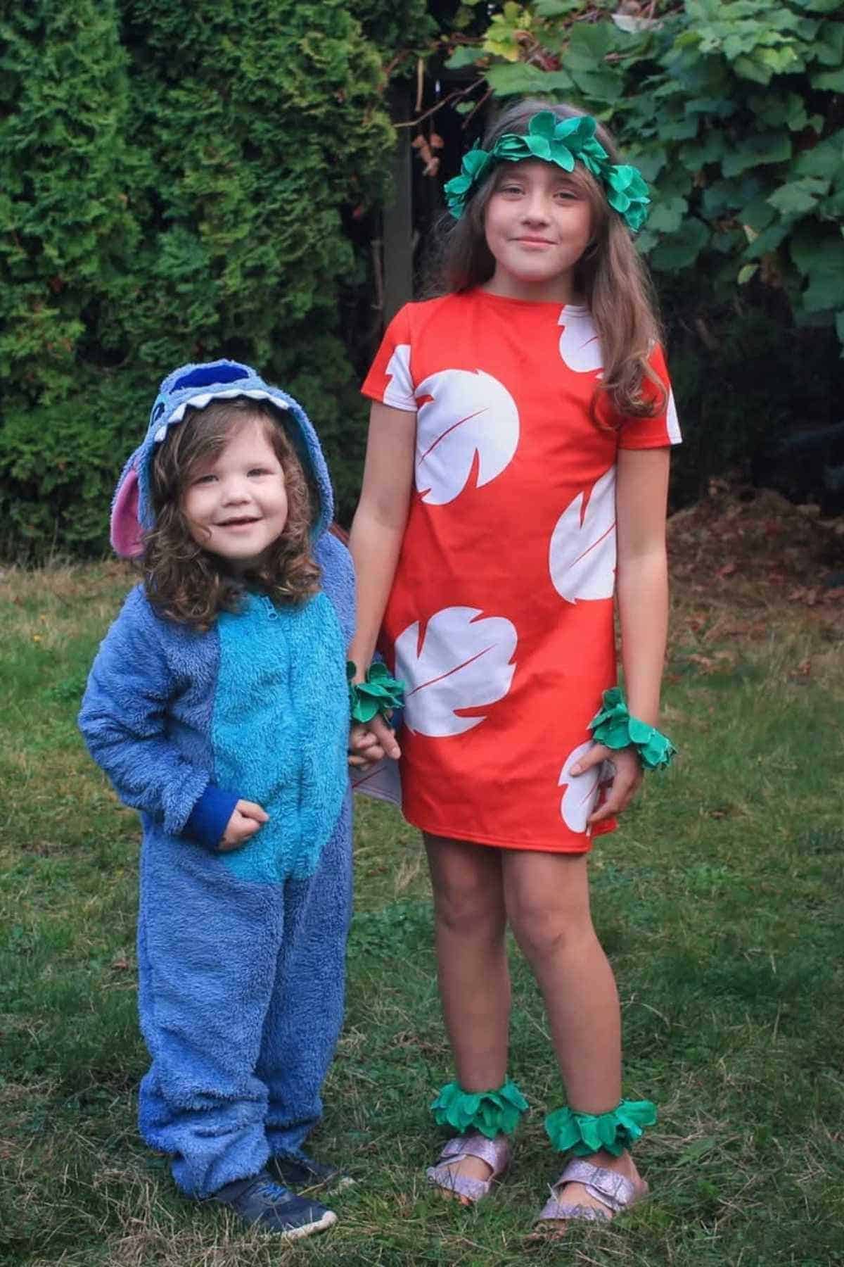 Two children standing on grass; one wears a blue furry costume with floppy ears, the other wears a red dress with white leaf patterns and green leafy accessories.