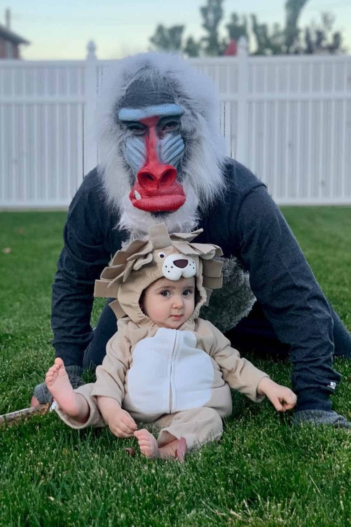 An adult wearing a baboon mask kneels on grass behind a toddler dressed in a lion costume, with a white fence and trees in the background.