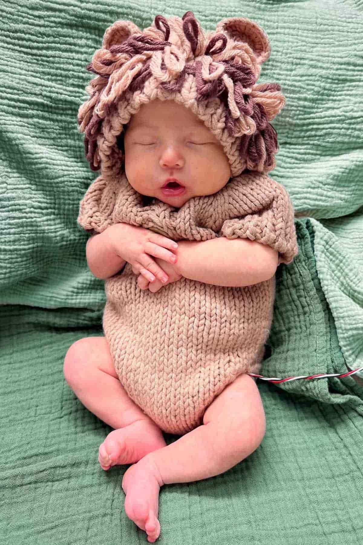 Baby wearing a knitted beige onesie and a matching hat with yarn loops, sitting with arms crossed and eyes closed on a green textured blanket.