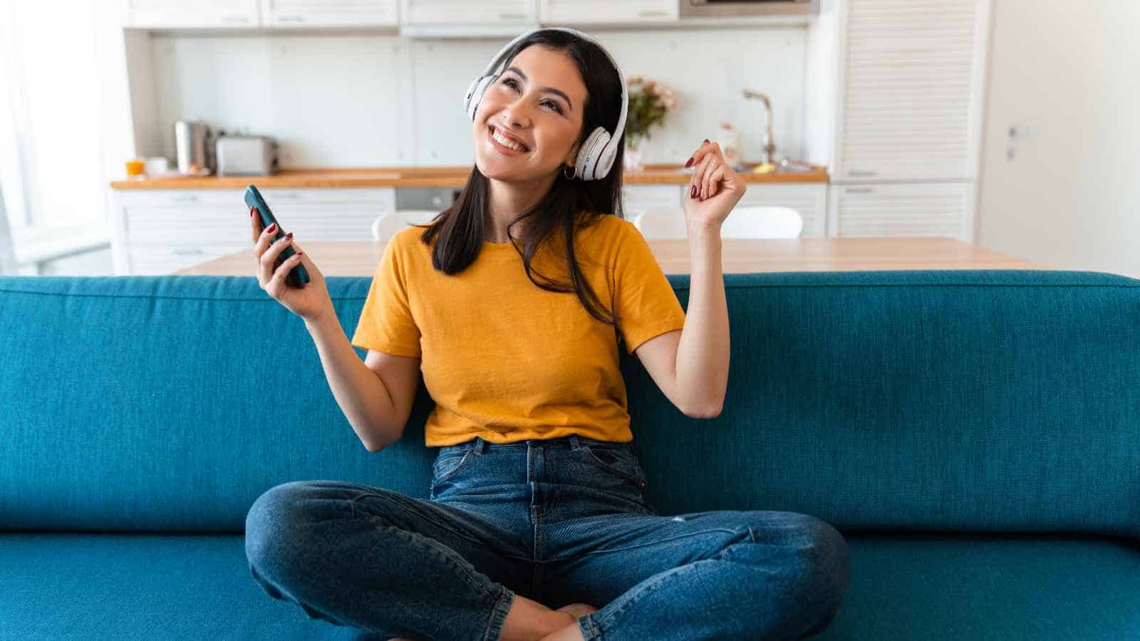 A woman sitting cross-legged on a blue sofa, wearing headphones and holding a smartphone, smiles and gestures with her hand.