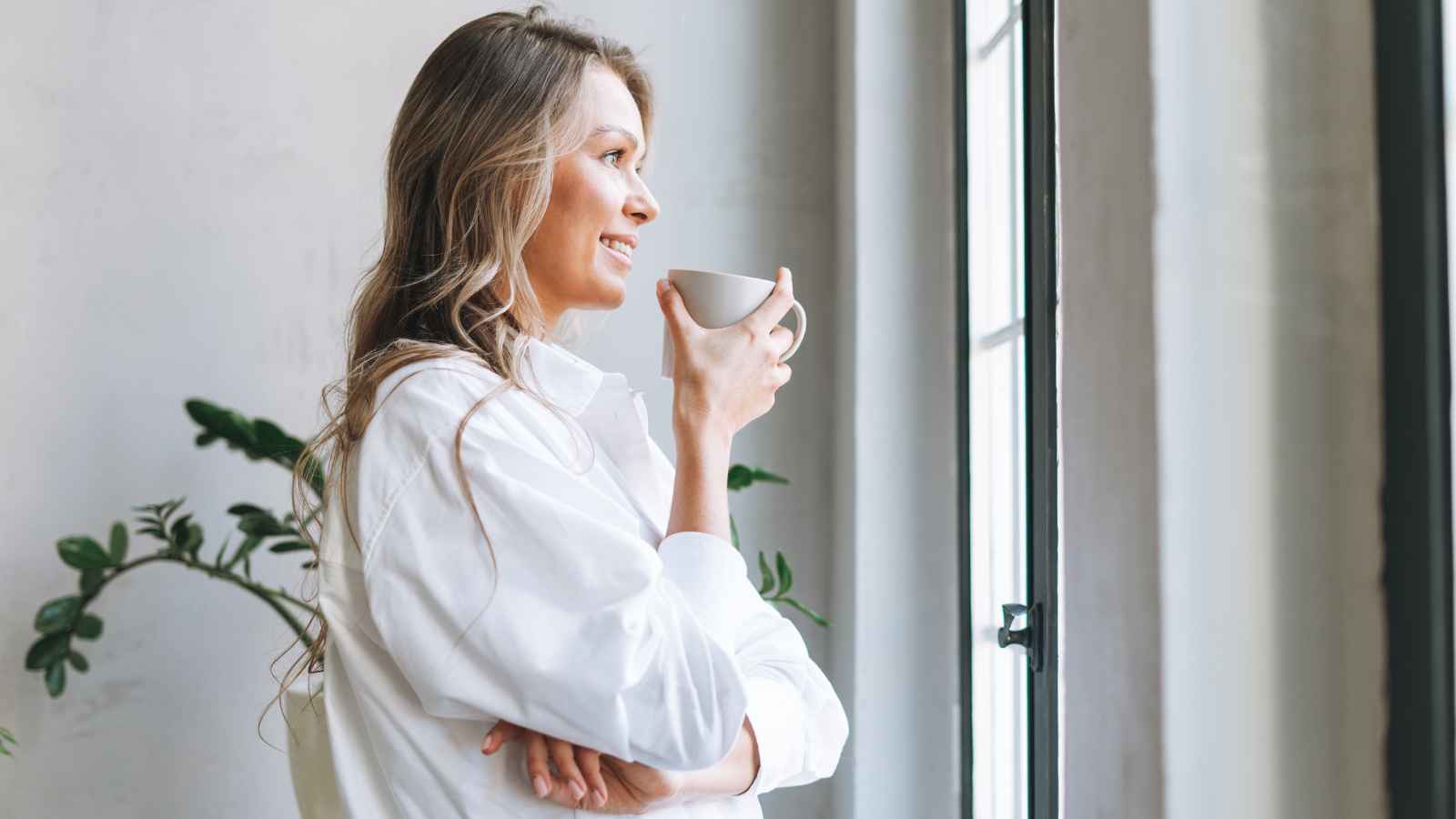 Woman in a white shirt holding a cup, standing by a window and looking outside with a slight smile. Indoor plant visible in the background.