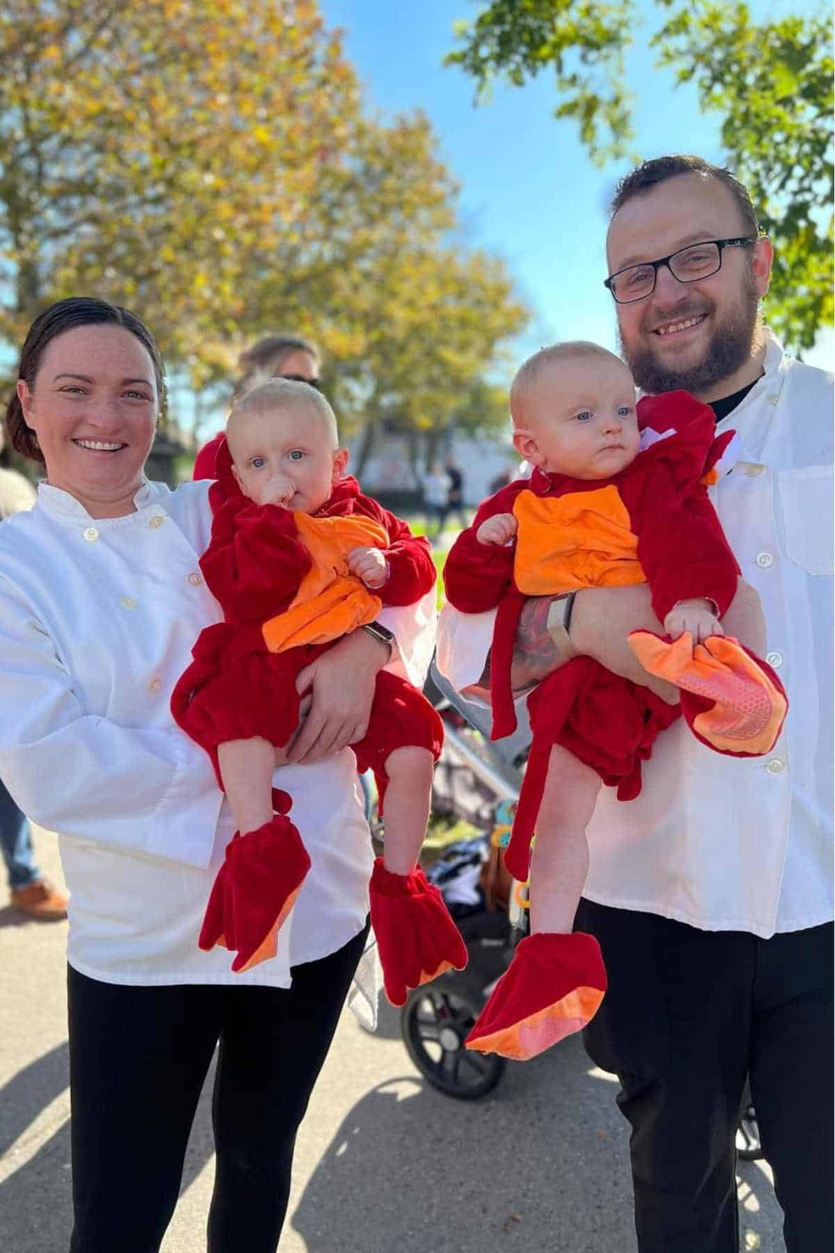 Two adults in white chef jackets each hold a baby dressed in a lobster costume outdoors on a sunny day.