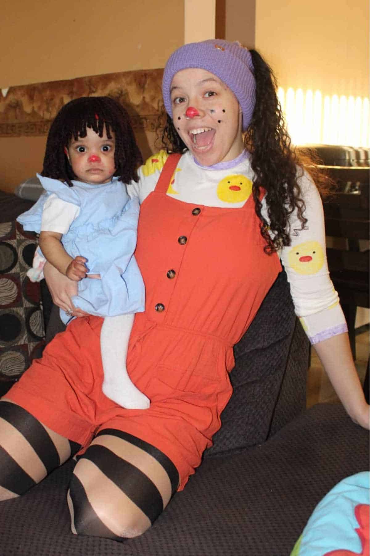 An adult and a child dressed in matching rag doll costumes, with painted faces, yarn wigs, and playful outfits, sitting on a couch.