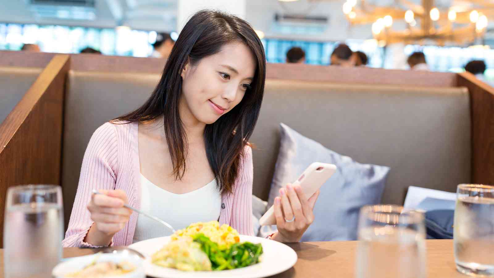 A woman sitting at a restaurant booth looks at her phone while eating a meal with pasta and vegetables.