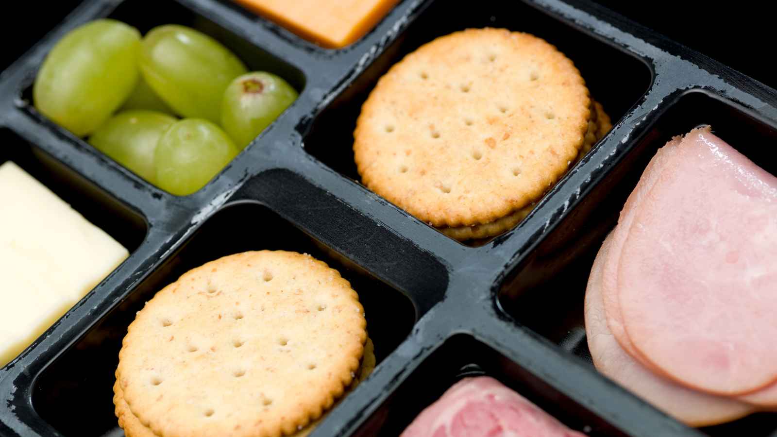 A close-up of a black tray containing round crackers, green grapes, cheese slices, and deli meat.