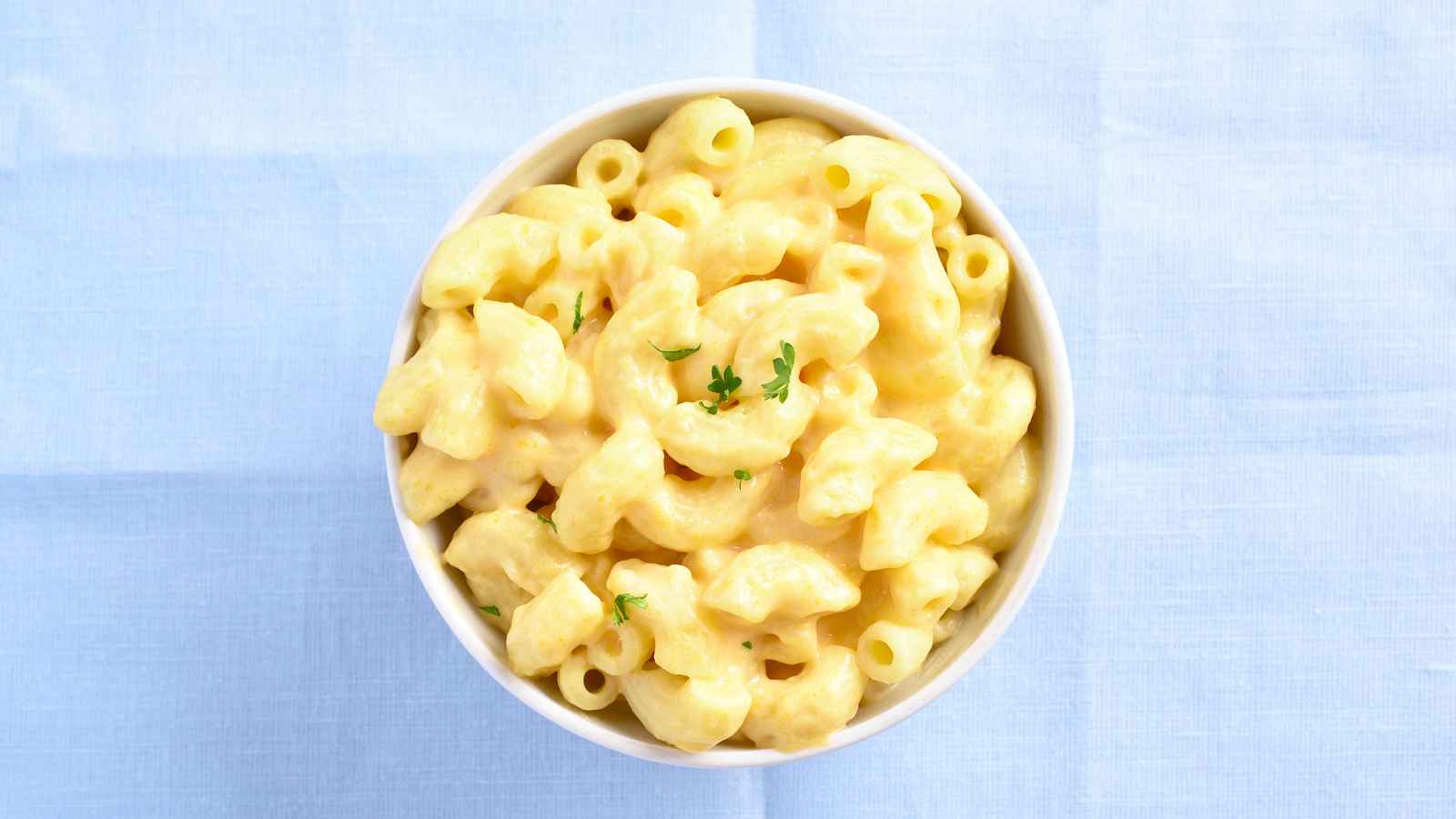A bowl of creamy macaroni and cheese topped with a few sprigs of parsley, placed on a light blue tablecloth.