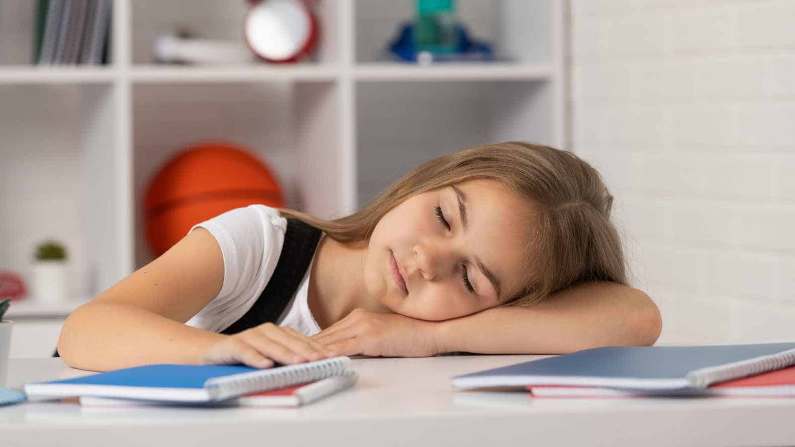 A young girl with long hair rests her head on her folded arm at a desk, eyes closed, with notebooks and shelves in the background.