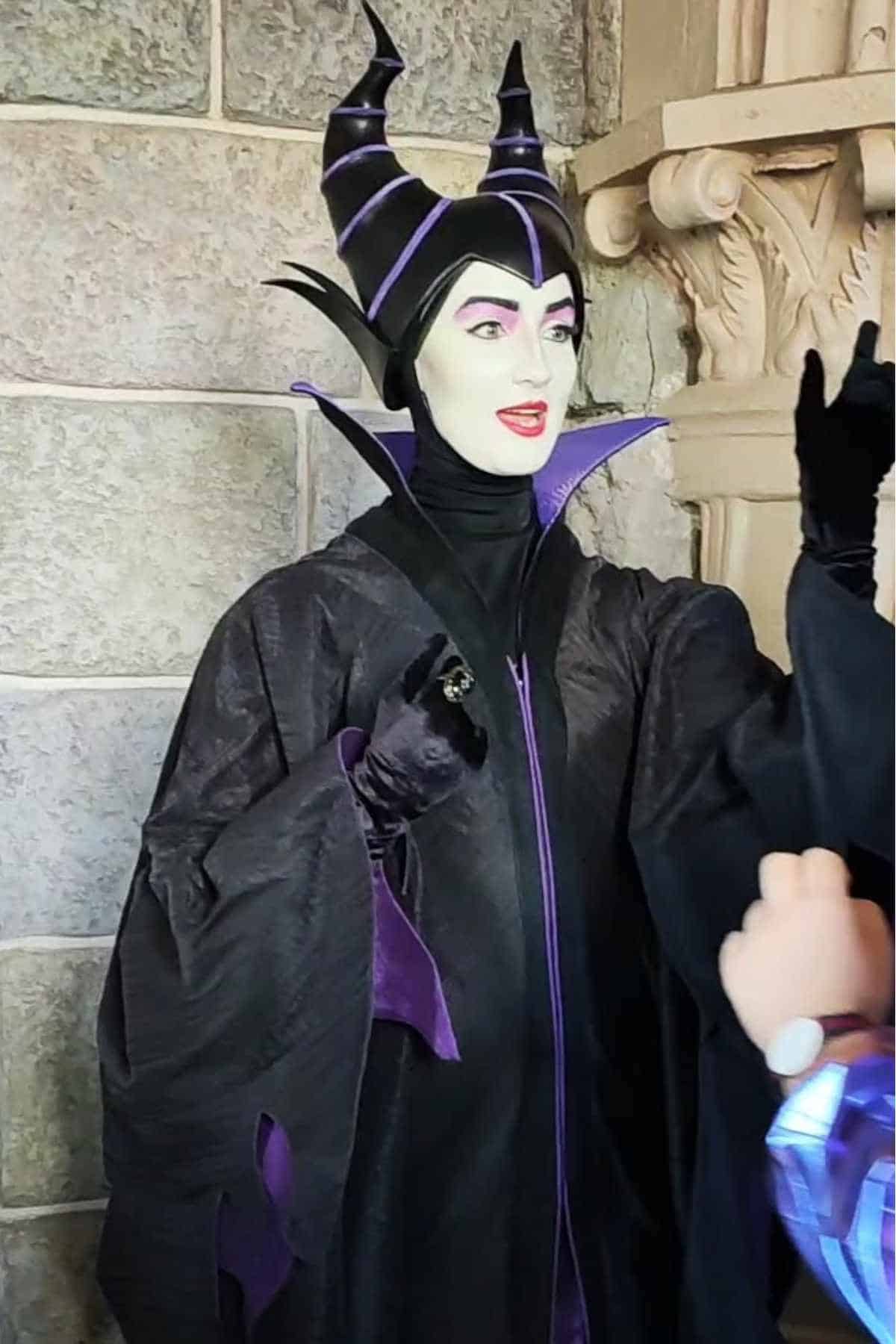 A person dressed as Maleficent, wearing a black and purple horned headpiece and robe, stands by a stone wall, gesturing with one hand raised.