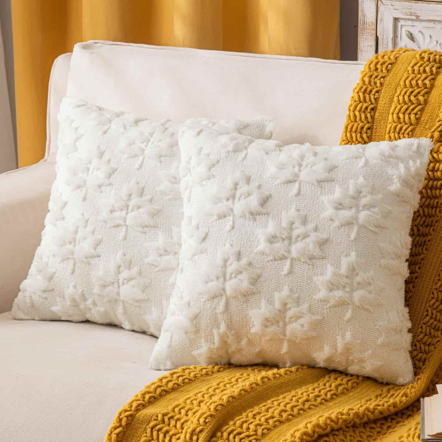 Two white textured throw pillows on a beige sofa, with a mustard yellow knitted blanket draped over the armrest.