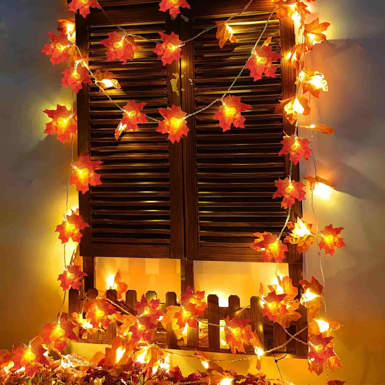 A window with closed wooden shutters is decorated with string lights featuring artificial autumn leaves, creating a warm, festive glow.