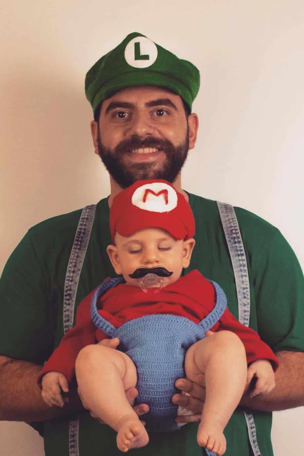 An adult dressed as Luigi holds a baby dressed as Mario, both wearing hats and costumes inspired by the Super Mario video game characters.