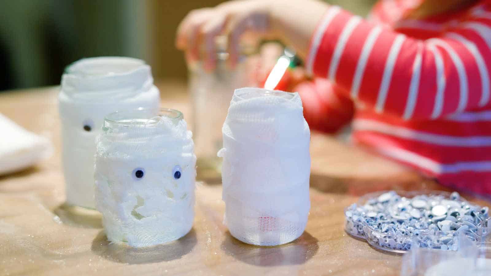 A child decorates glass jars wrapped in white gauze with googly eyes, crafting them to resemble mummies on a table.