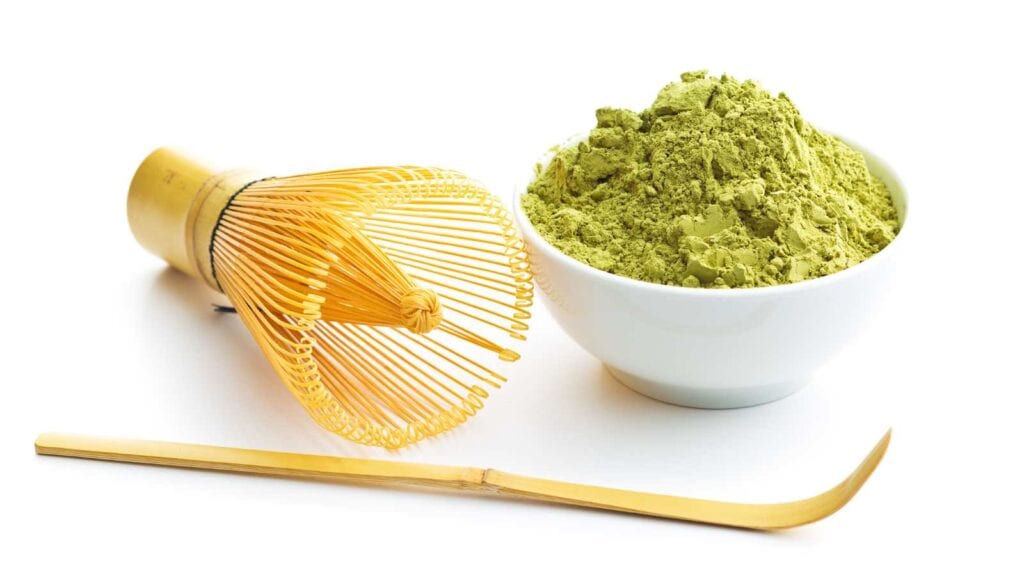 A bamboo whisk, a bamboo scoop, and a white bowl filled with green matcha powder are arranged on a white surface.