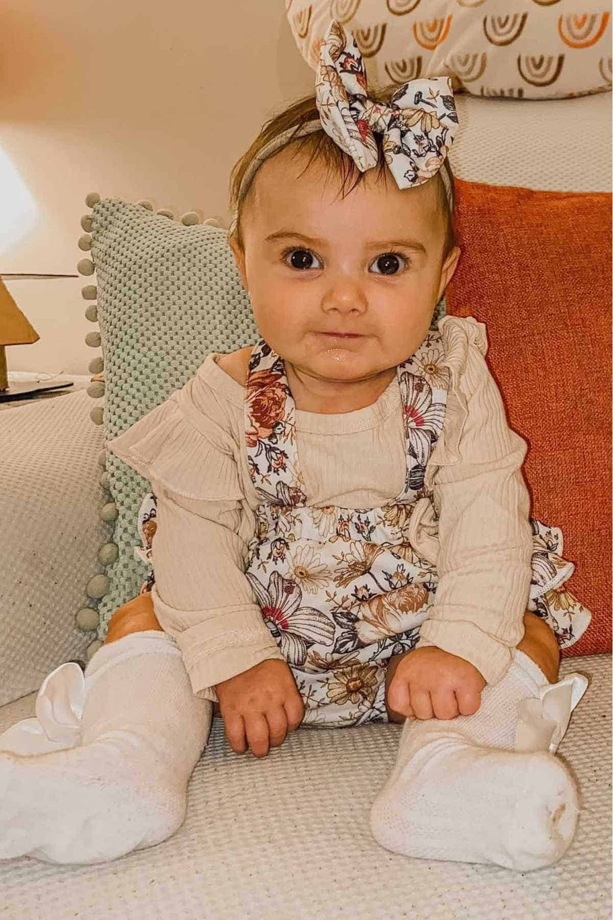 A baby wearing a floral headband and matching dress sits on a couch with textured pillows, looking directly at the camera.