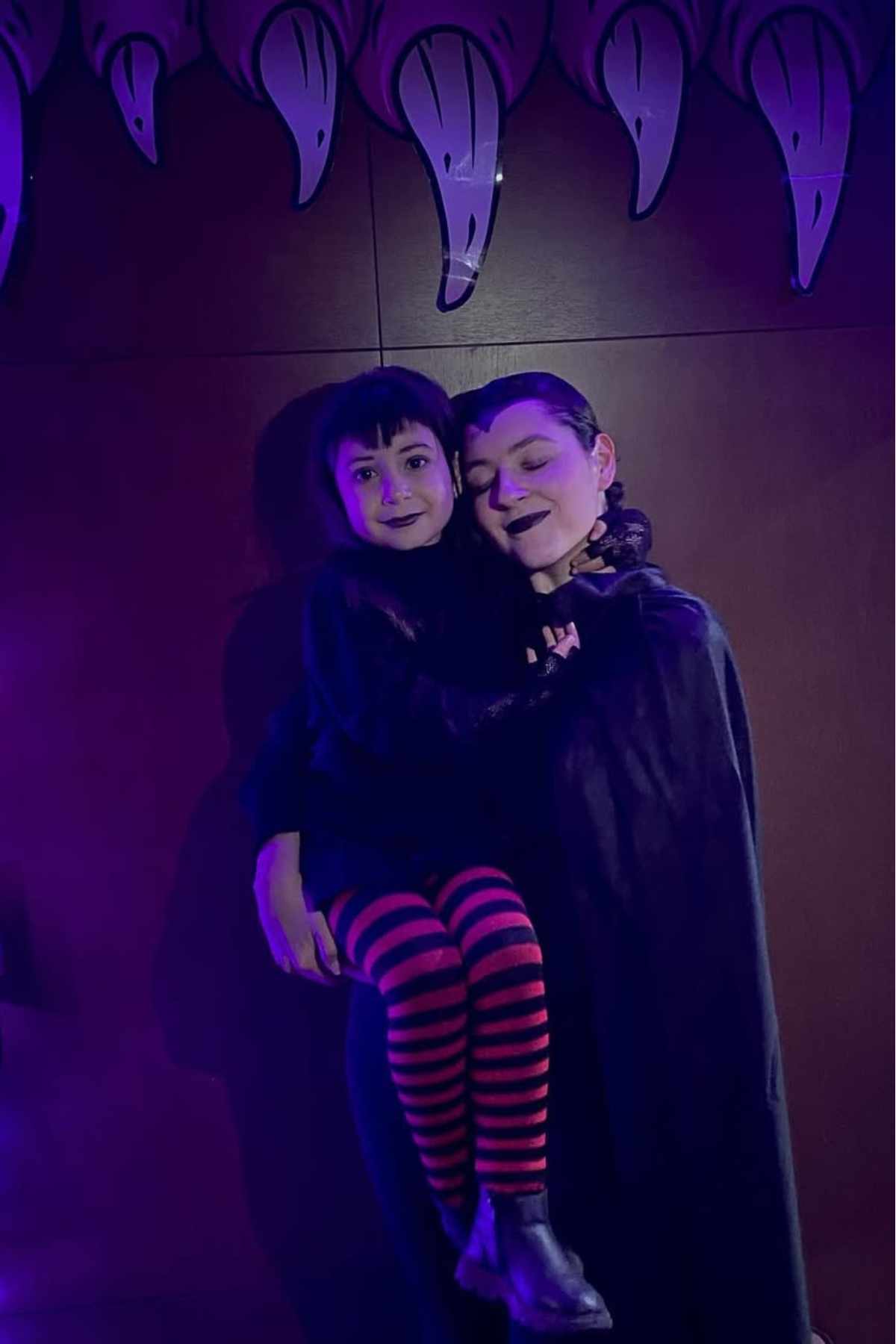 An adult and a child in dark clothing and makeup pose together under pointed decorations in a dim, purple-lit room.