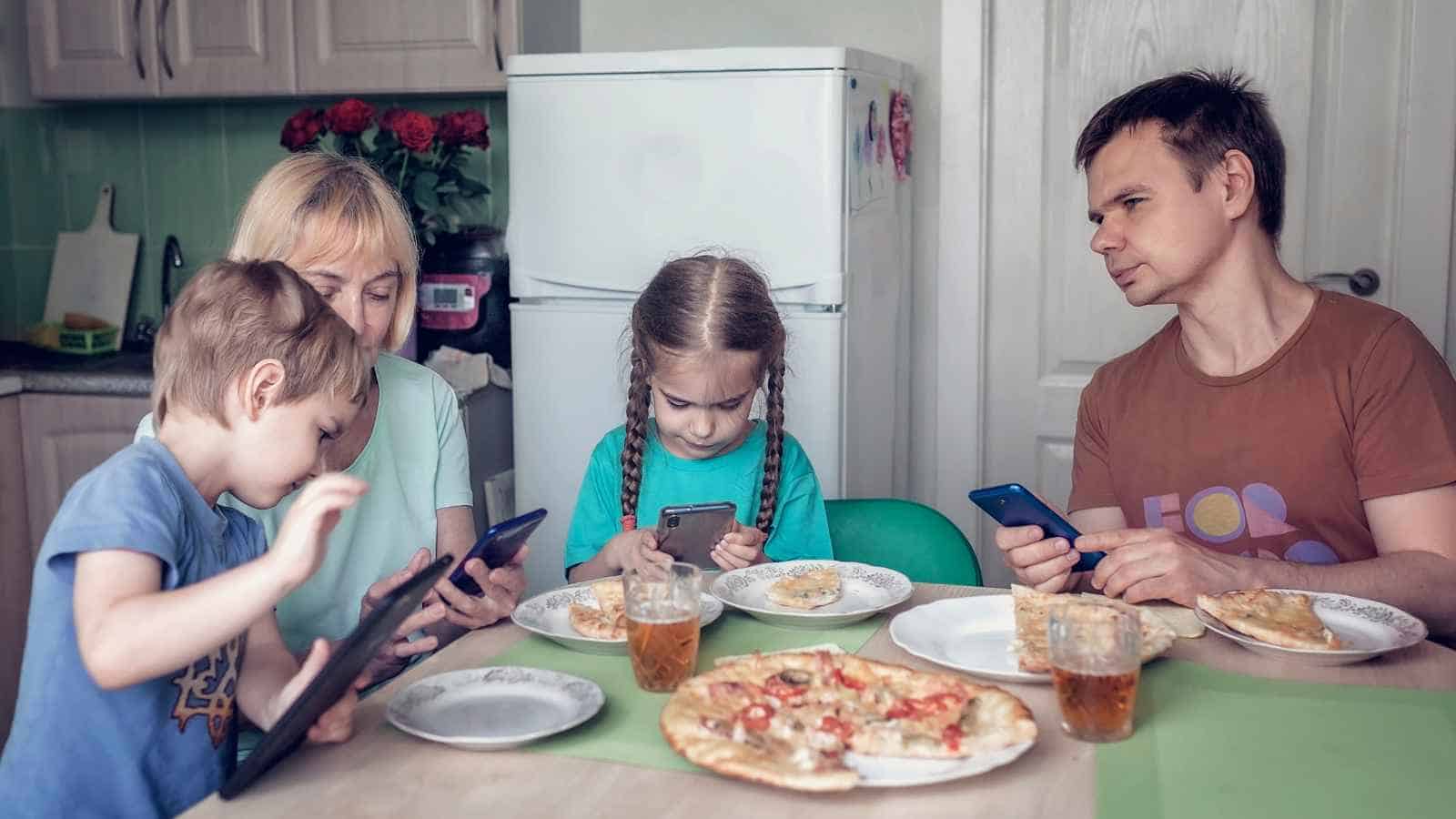A family of four sits at a table with pizza and drinks, each person focused on their own electronic device.
