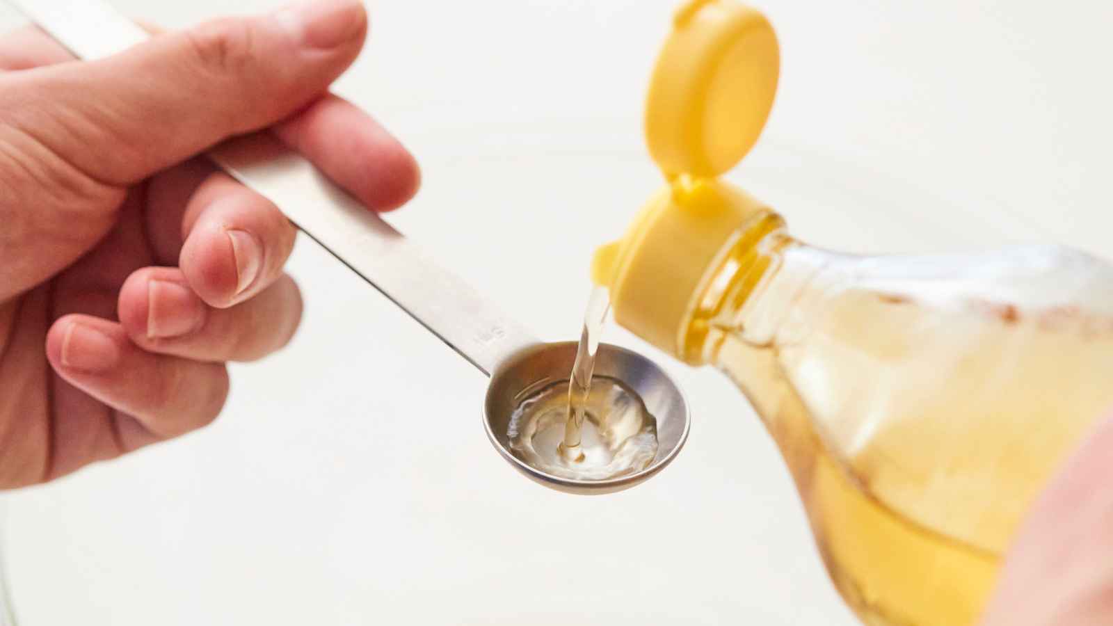 A person pours oil from a plastic bottle into a metal tablespoon held above a bowl.