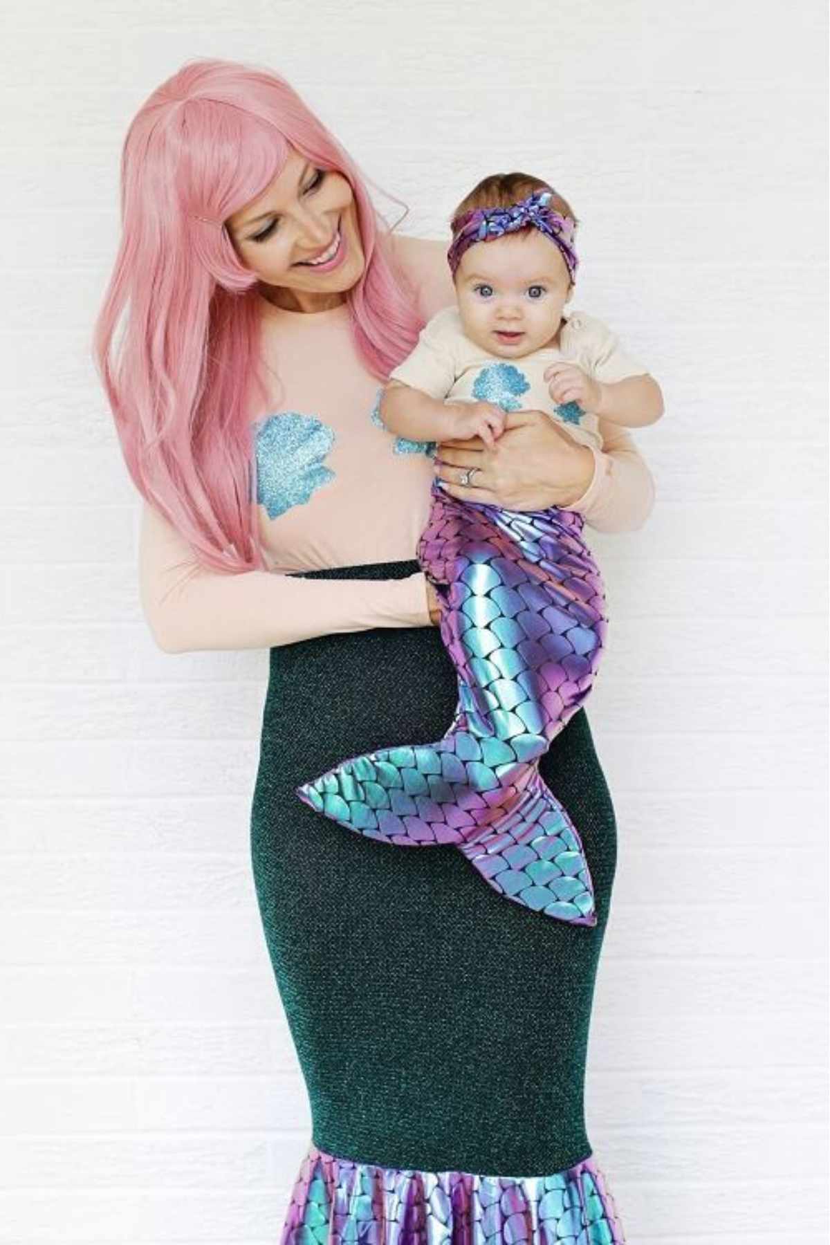 A woman in a pink wig and mermaid costume holds a baby dressed in a matching mermaid tail and headband against a white background.