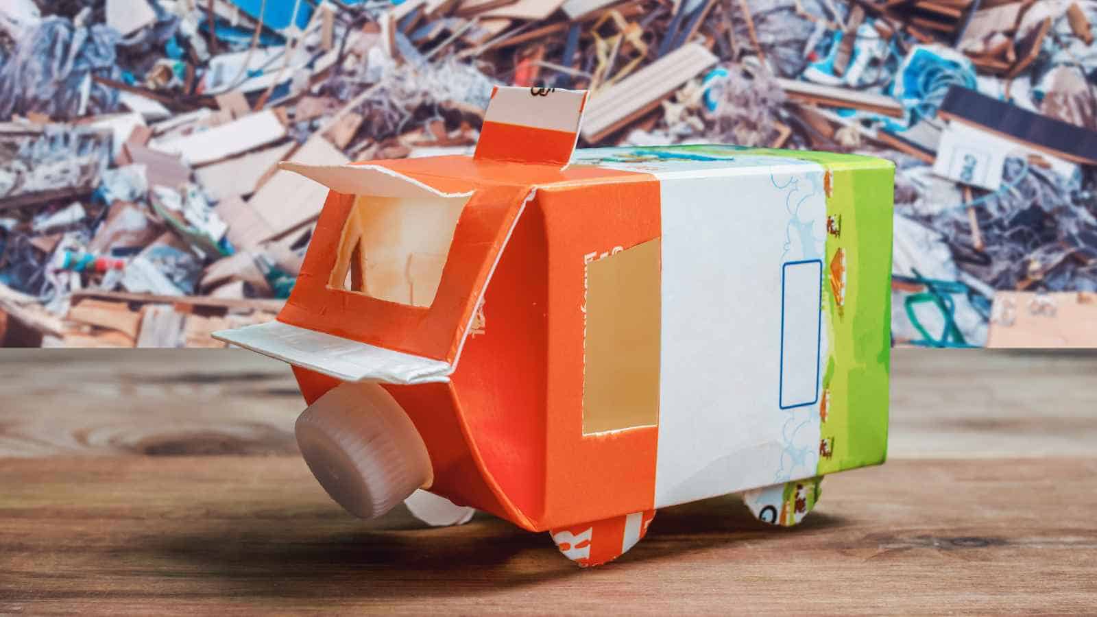 A small vehicle model made from recycled cartons and a plastic bottle cap, placed on a wooden surface with a pile of junk in the background.