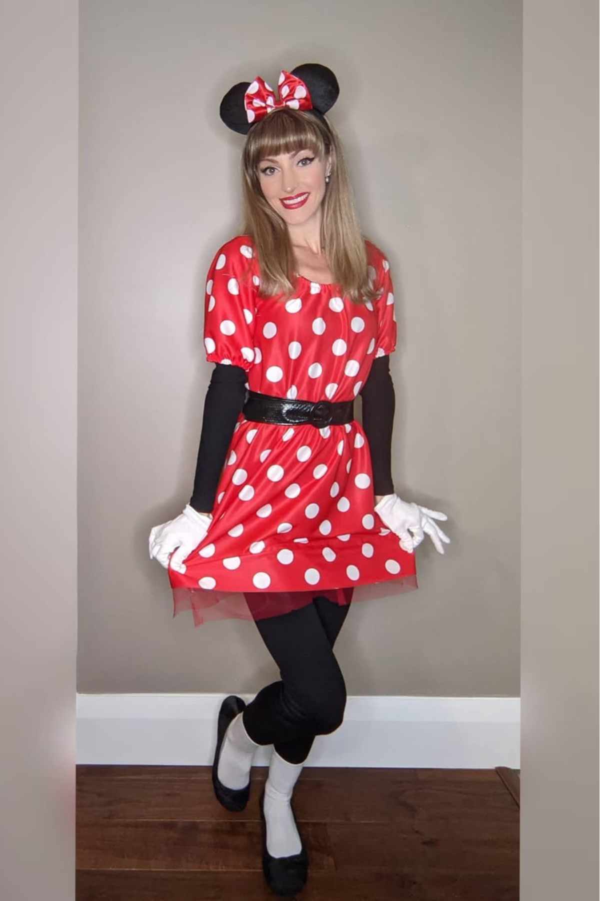 Person dressed in a Minnie Mouse costume, featuring a red polka dot dress, black tights, white gloves, mouse ears headband with a bow, standing against a plain wall.