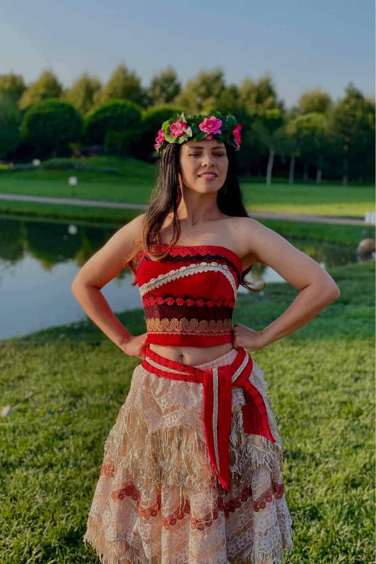 A woman stands outdoors near water, wearing a floral crown, a red and brown patterned crop top, and a layered beige skirt with red accents.