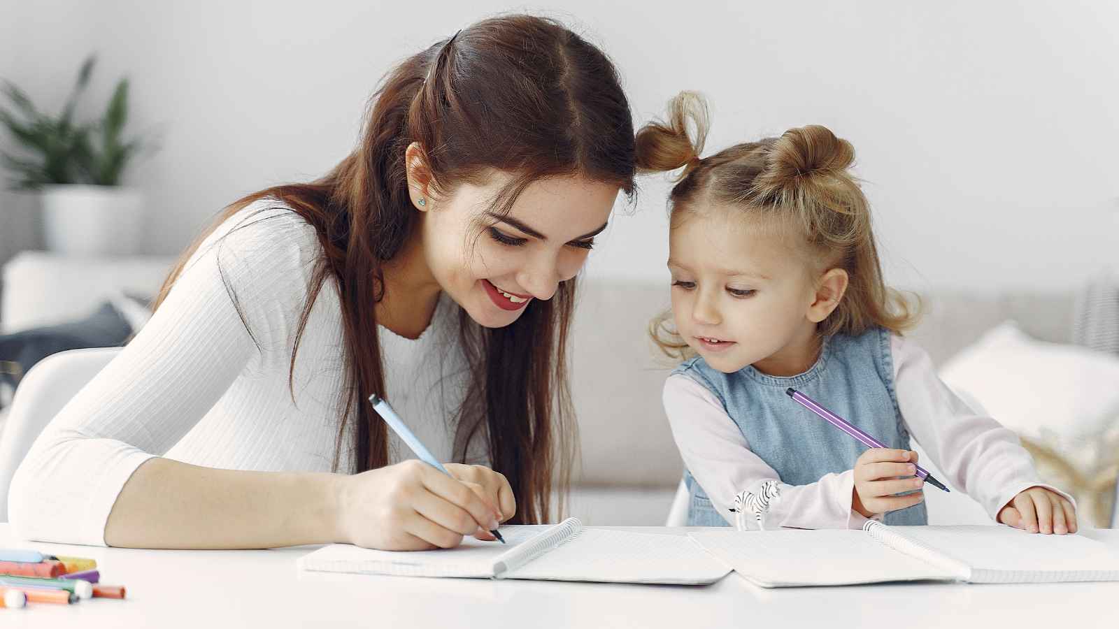 A woman and a young girl sit at a table, smiling and writing in notebooks together. Colored pencils are scattered nearby.