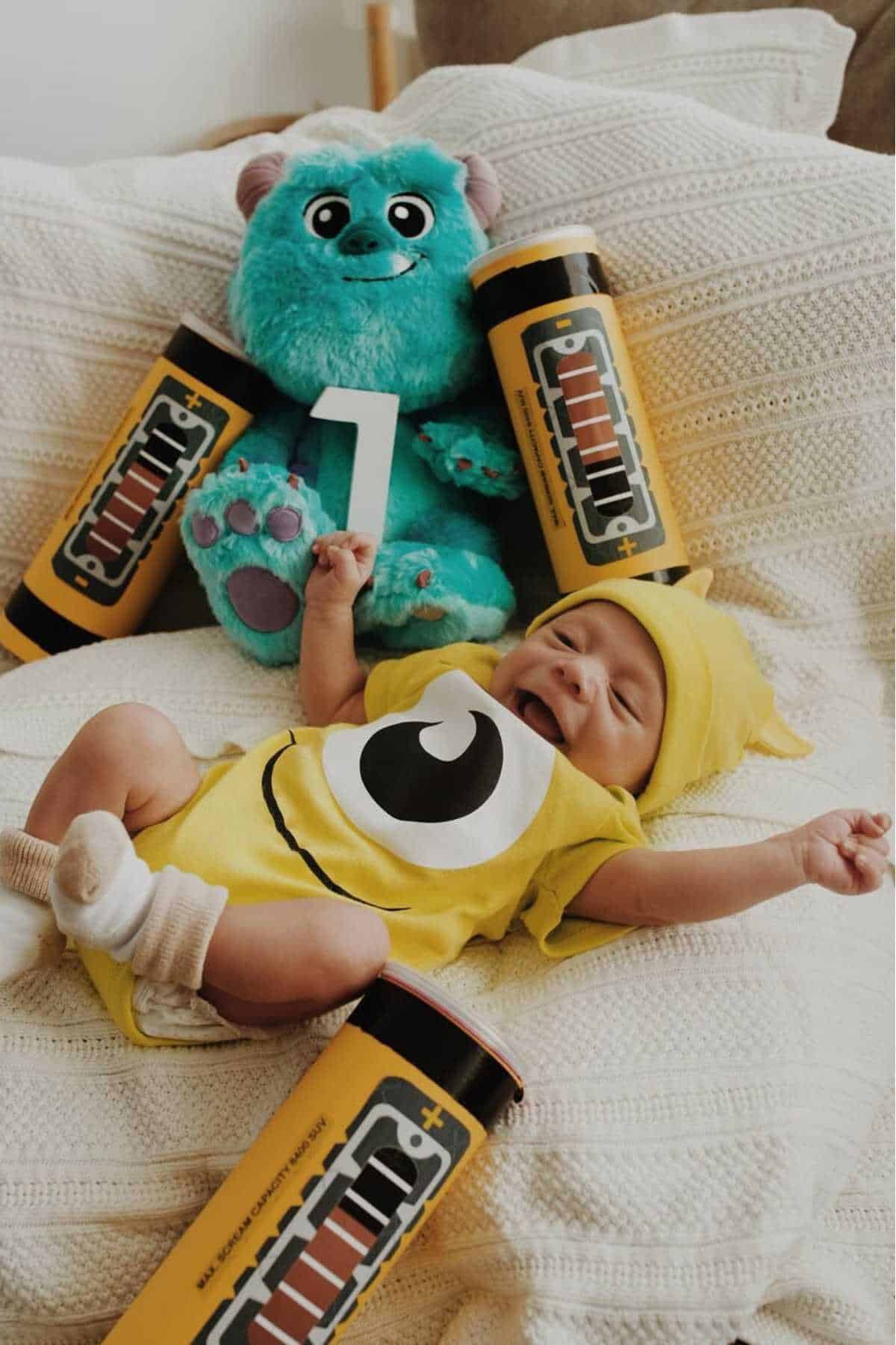 A baby in a yellow monster costume lies on a blanket, surrounded by large battery props and a blue plush monster toy holding a number one.