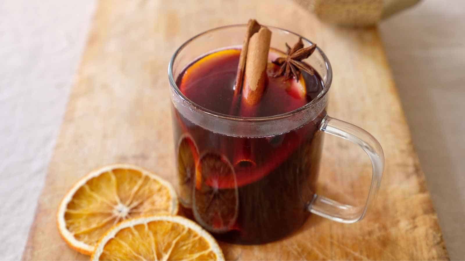 A glass mug of mulled wine with orange slices, a cinnamon stick, and star anise on a wooden surface, next to two dried orange slices.