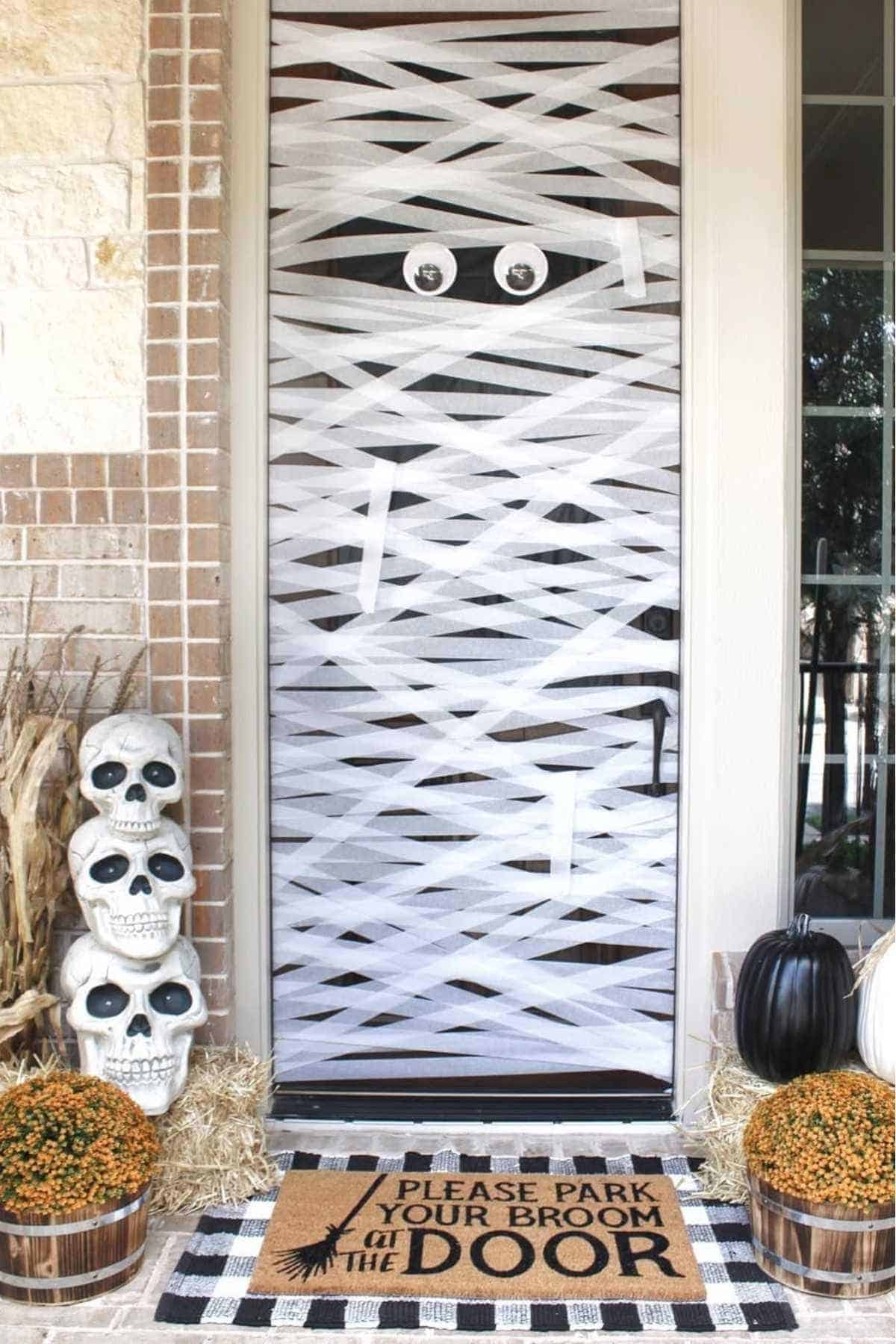 A front door decorated like a mummy with white streamers and googly eyes, next to stacked skulls, pumpkins, and a mat that says, "Please park your broom at the door".