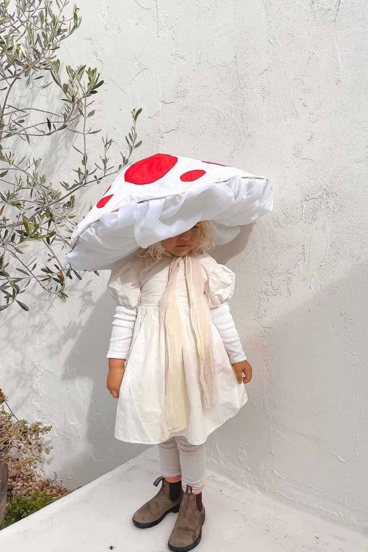 A young child wearing a large mushroom cap costume with red spots, white dress, white leggings, and brown boots, standing against a plain white wall.