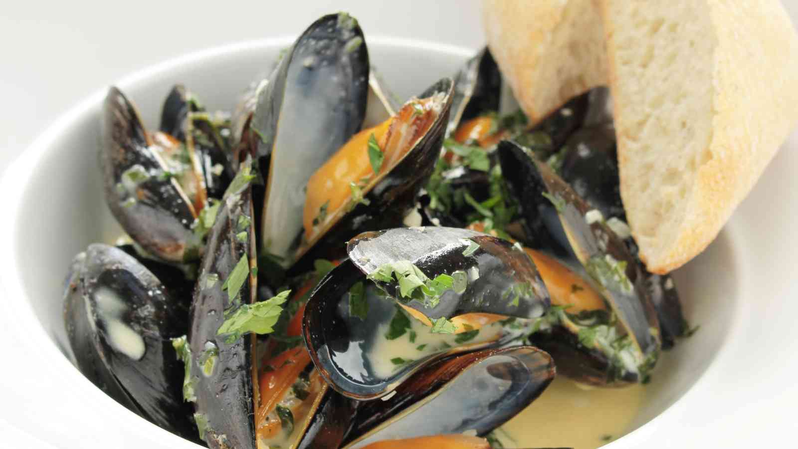 A bowl of steamed mussels in a creamy herb sauce, garnished with chopped parsley, served with slices of crusty bread.