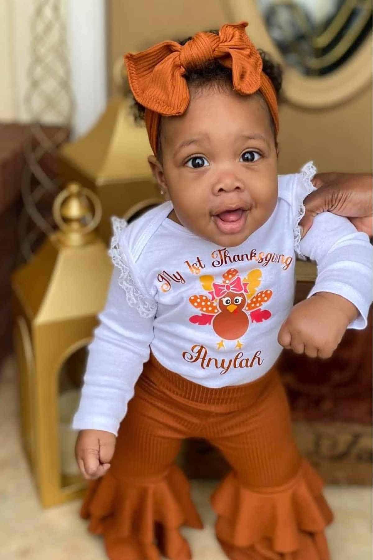 A baby wearing a headband and an outfit that reads "My 1st Thanksgiving Anylah" stands while being supported by an adult hand.