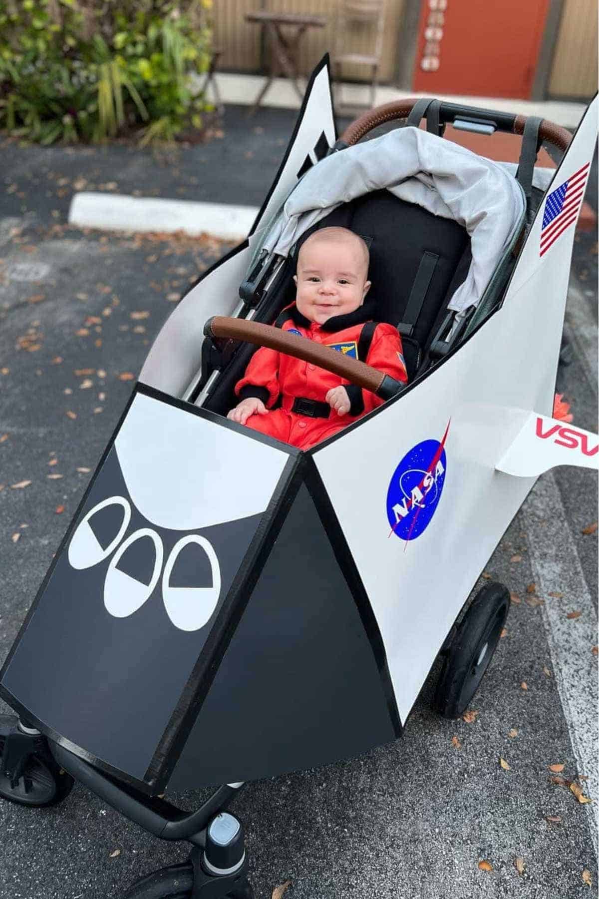 A baby in a red astronaut suit sits in a stroller decorated to look like a NASA space shuttle, outdoors in a parking lot.