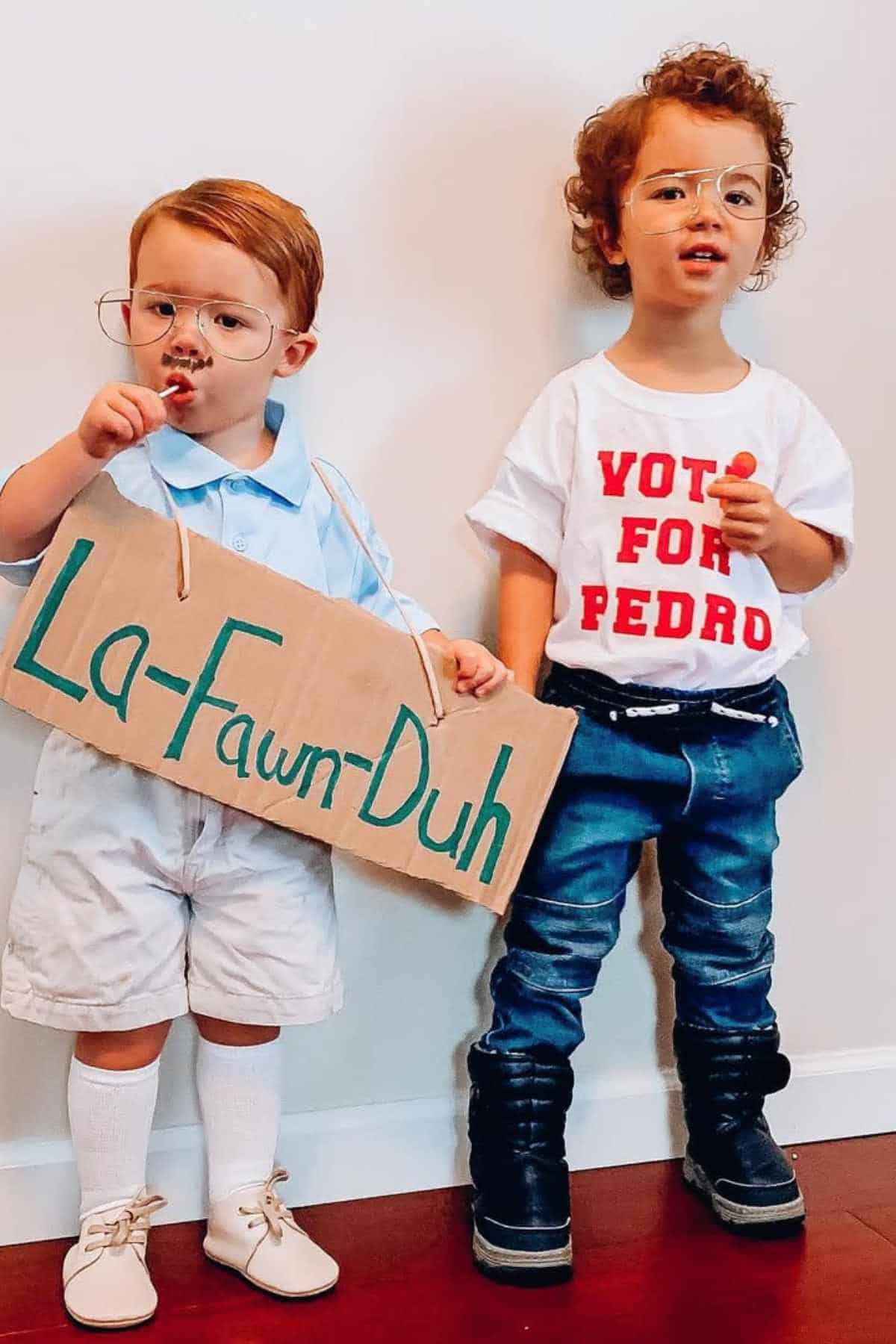 Two young children in costume stand against a wall. One holds a "La-Fawn-Duh" sign, the other wears a "Vote for Pedro" shirt. Both wear glasses and hold lollipops.